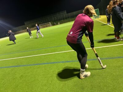 A girl with her hockey stick on the ground looking at a girl in the distance on the pitch